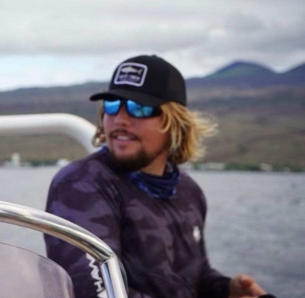 Man with sunglasses and cap smiling on a boat with mountains in the background.