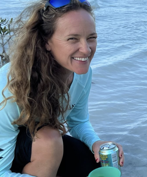 Smiling person kneeling by water, holding a drink can, wearing a light blue top.