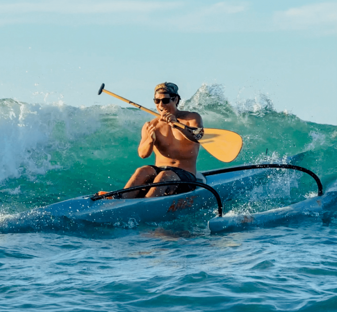 Man in a kayak paddling on a wave in the ocean, wearing sunglasses and a cap.
